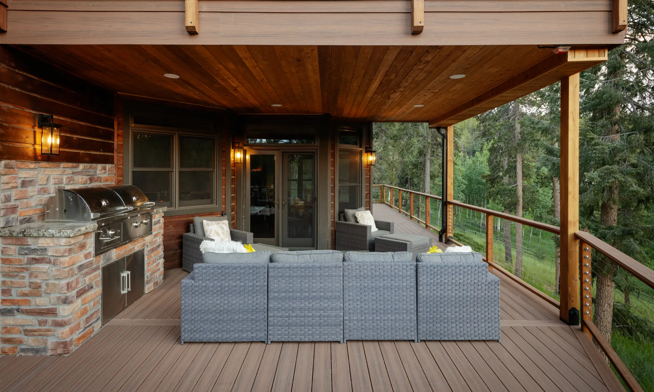 Covered deck with a warm wood ceiling, wicker sectional, built-in grill in a stone surround and cable railing facing the trees