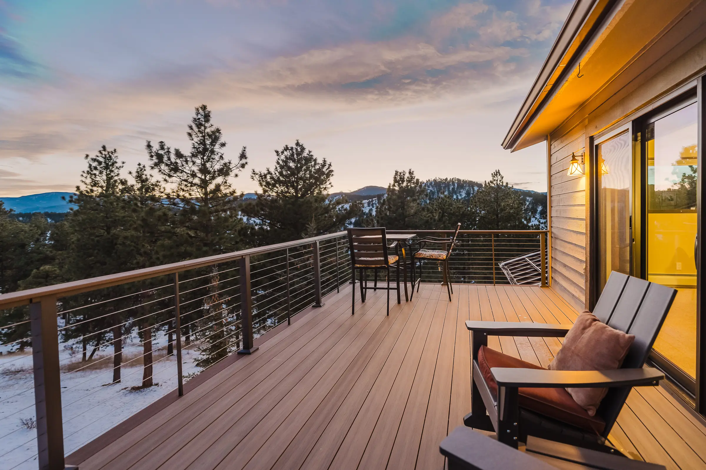 Deck with cable railing and lounge chairs at sunset, looking out over pines and snowy mountains